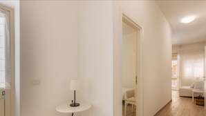 A hallway with a round table and a lamp, leading to a room with a bed and a chair. - Anita Apartment (Noto)