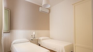 A bedroom with two single beds, a bedside table with a lamp, a wall-mounted air conditioner, and a window with blinds. - Anita Apartment (Noto)