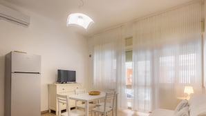 A compact room with a bed, a desk with a television, a refrigerator, and a window with sheer curtains. - Anita Apartment (Noto)