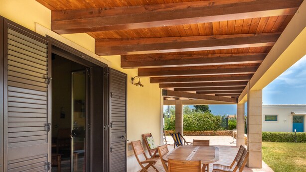 An outdoor patio with a wooden table and chairs, a covered area with a wooden ceiling, and a view of a building in the background. - Garden Chich (Noto)