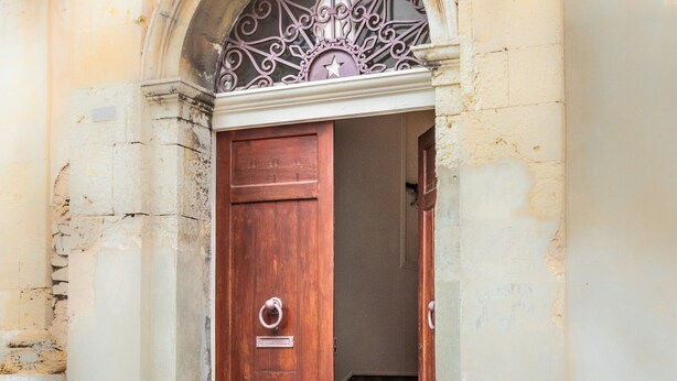 A wooden door with a metal handle and a nameplate, set in a stone building with a decorative metal arch above it. - Liberty Guest House (Avola)