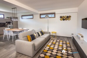 A modern living room with a sofa, dining table, and chairs. There is a kitchen area in the background with a stove and cabinets. - Magnolia (Avola)