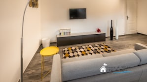 A modern living room with a sofa, a yellow side table, a patterned rug, a TV mounted on the wall, and a cabinet with books.