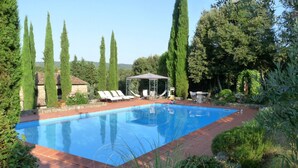 A rectangular swimming pool with a tiled edge, surrounded by a brick patio. There are lounge chairs and a covered seating area with a metal frame. Tall, slender trees are visible in the background.