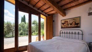 A bedroom with a large bed, wooden ceiling beams, a painting on the wall, and a view of trees and a gravel path through open doors.