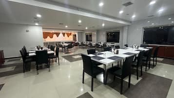 Una amplia sala de conferencias con varias mesas y sillas, una zona de cocina al fondo y grandes ventanales que proporcionan luz natural.