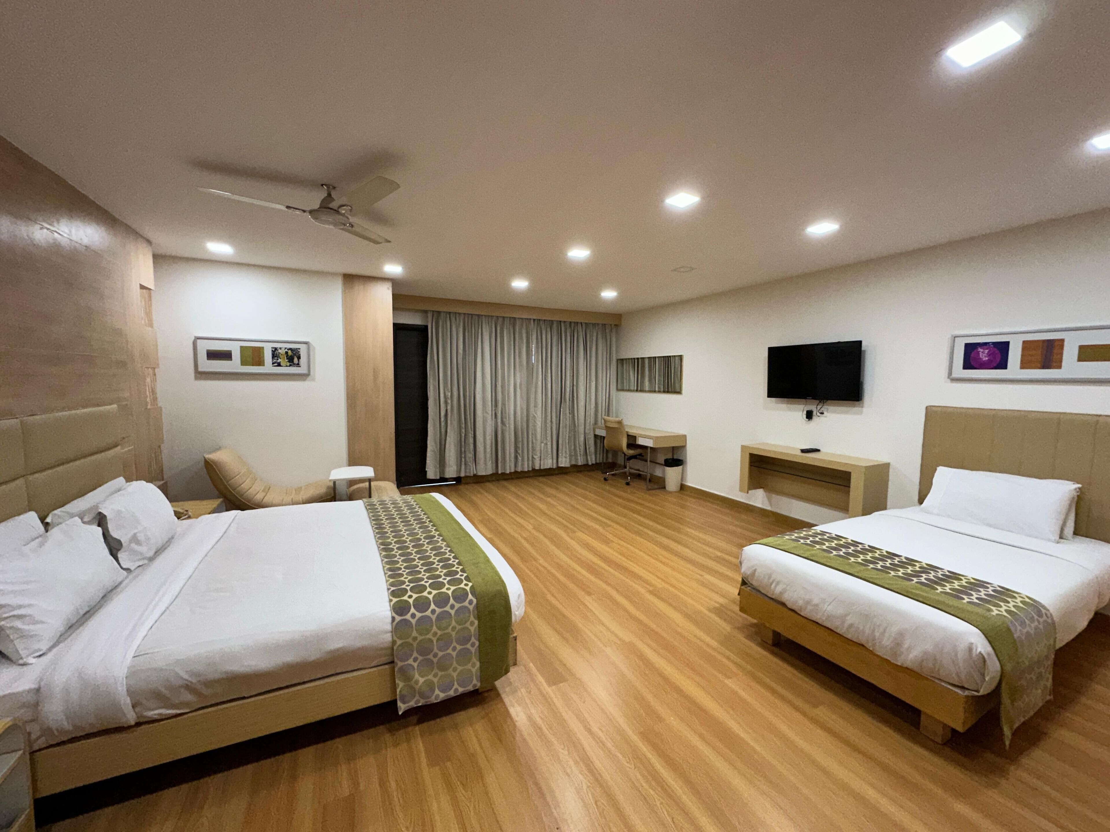 A hotel room with two beds, a desk, a chair, a television, and a ceiling fan.