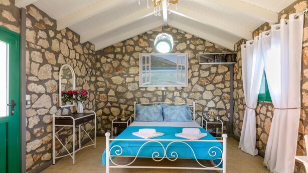 Une chambre avec un mur en pierre, un lit recouvert d’une courtepointe bleue, un lustre et une vue sur la mer par la fenêtre.