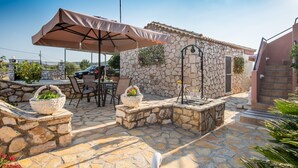 Un espace extérieur aménagé en terrasse, avec un sol en pierre, une table entourée de chaises, un grand parasol et un mur en pierre doté d’une porte.