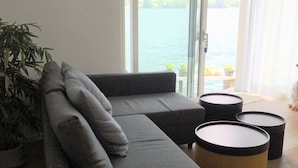 Una sala de estar moderna con un sofá seccional gris, una planta en maceta y vista a una masa de agua a través de ventanas amplias.