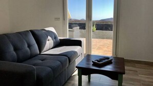 A living room with a dark grey sofa, a wooden coffee table, and a sliding glass door leading to a patio with a view of mountains.
