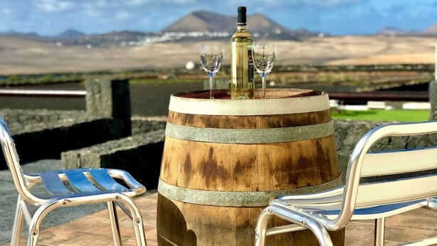 A wooden barrel table with a bottle and glasses, two metal chairs, and a view of a mountain in the background.