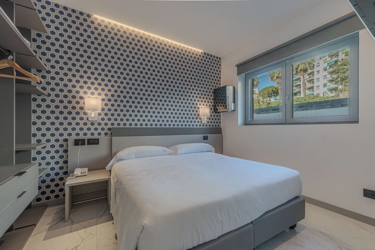 A modern bedroom with a geometric patterned wall, a bed with white bedding, bedside tables, a wardrobe, and a window with a view of buildings.