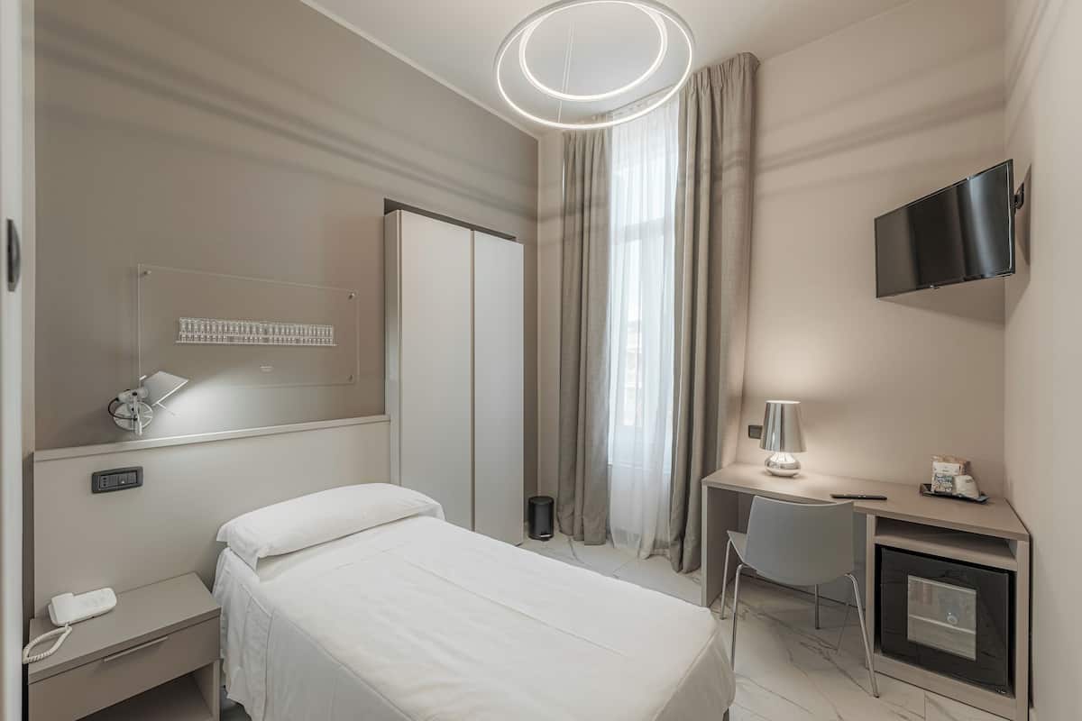 A hotel room with a bed, bedside table, desk with a lamp, a TV mounted on the wall, and a window with curtains.