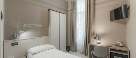 A hotel room with a bed, bedside table, desk with a lamp, a TV mounted on the wall, and a window with curtains.
