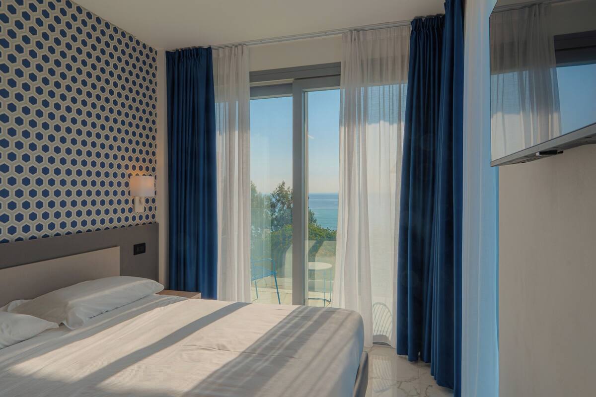 A hotel room with a bed, a patterned wall, a view of the outdoors, and a TV on the wall.