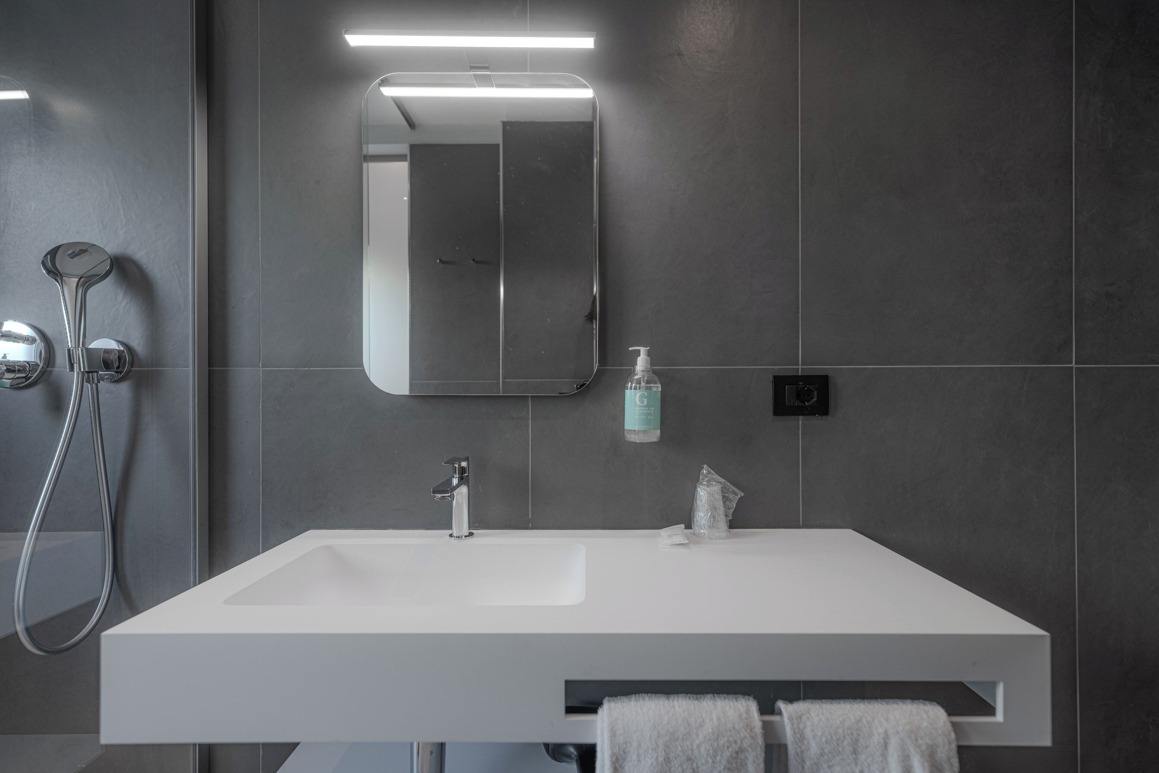 Un baño moderno con un lavabo blanco, un espejo, un cabezal de ducha de mano y un dispensador de jabón.