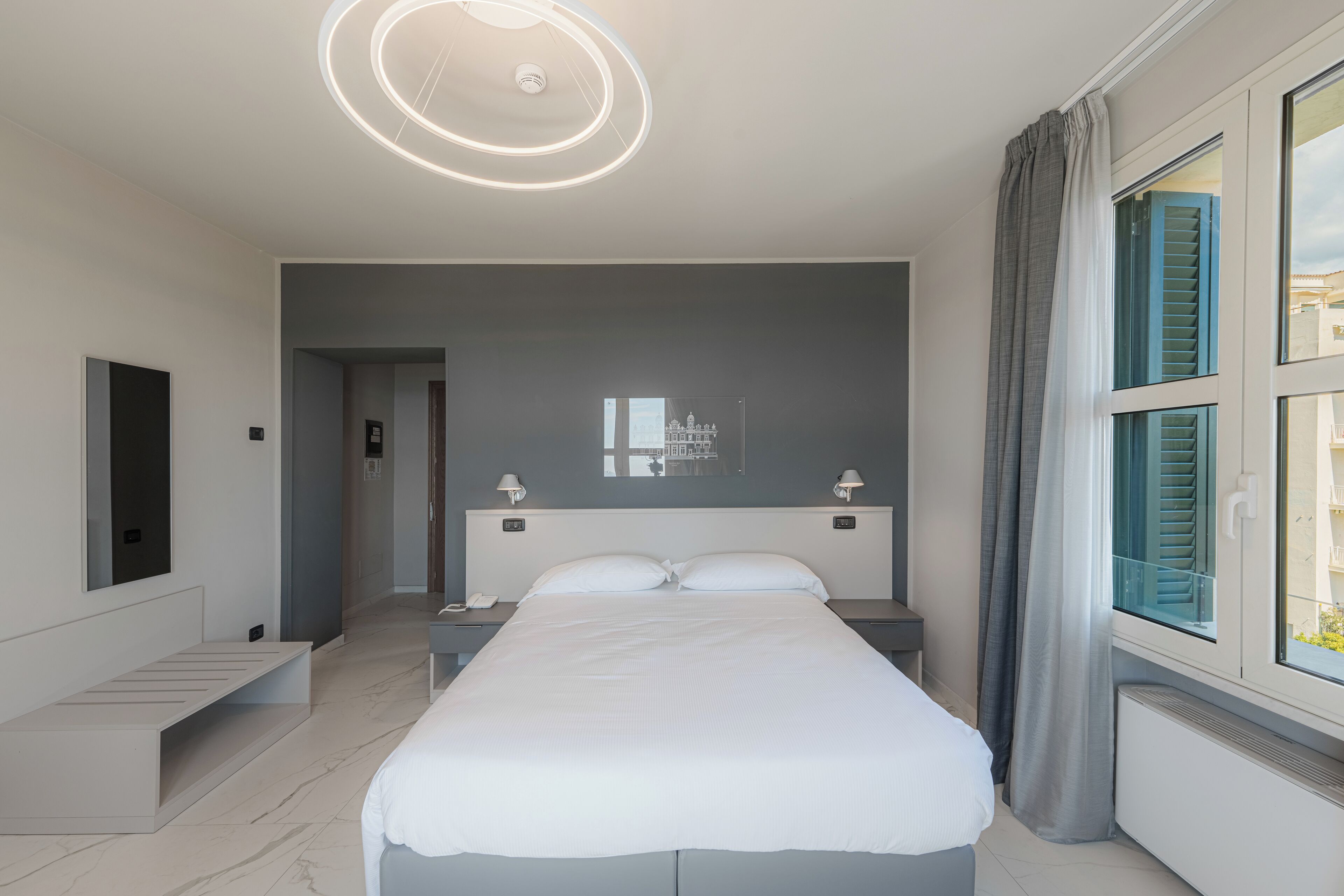 A modern hotel room with a large bed, bedside tables, a built-in bench, a ceiling light fixture, and a window with curtains.
