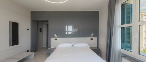 A modern hotel room with a large bed, bedside tables, a built-in bench, a ceiling light fixture, and a window with curtains.