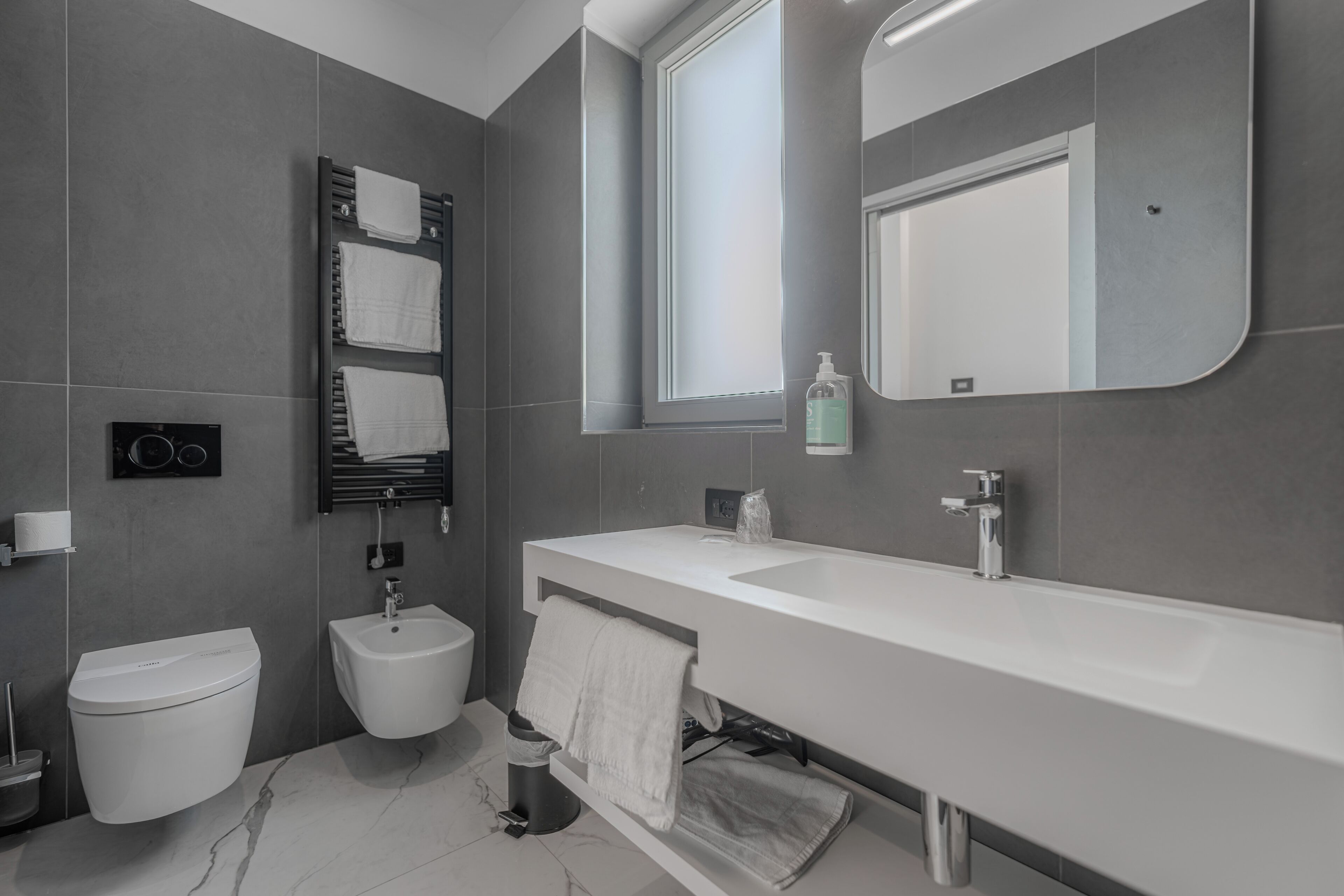 A modern bathroom with a white sink, bidet, and toilet, a towel rack, and a mirror.