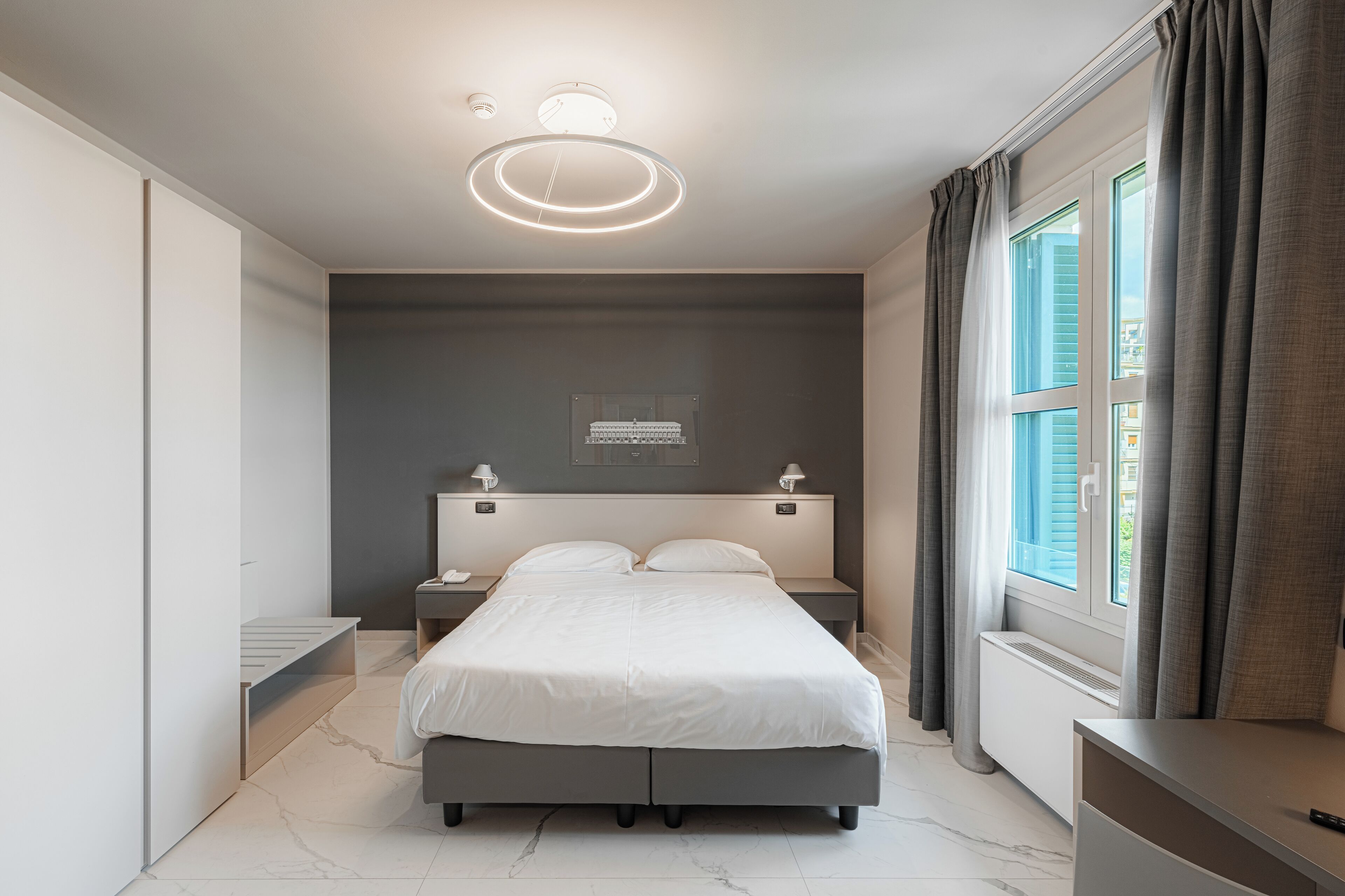Une chambre moderne avec un grand lit, des tables de chevet, un banc intégré, un luminaire au plafond et une fenêtre avec des rideaux.