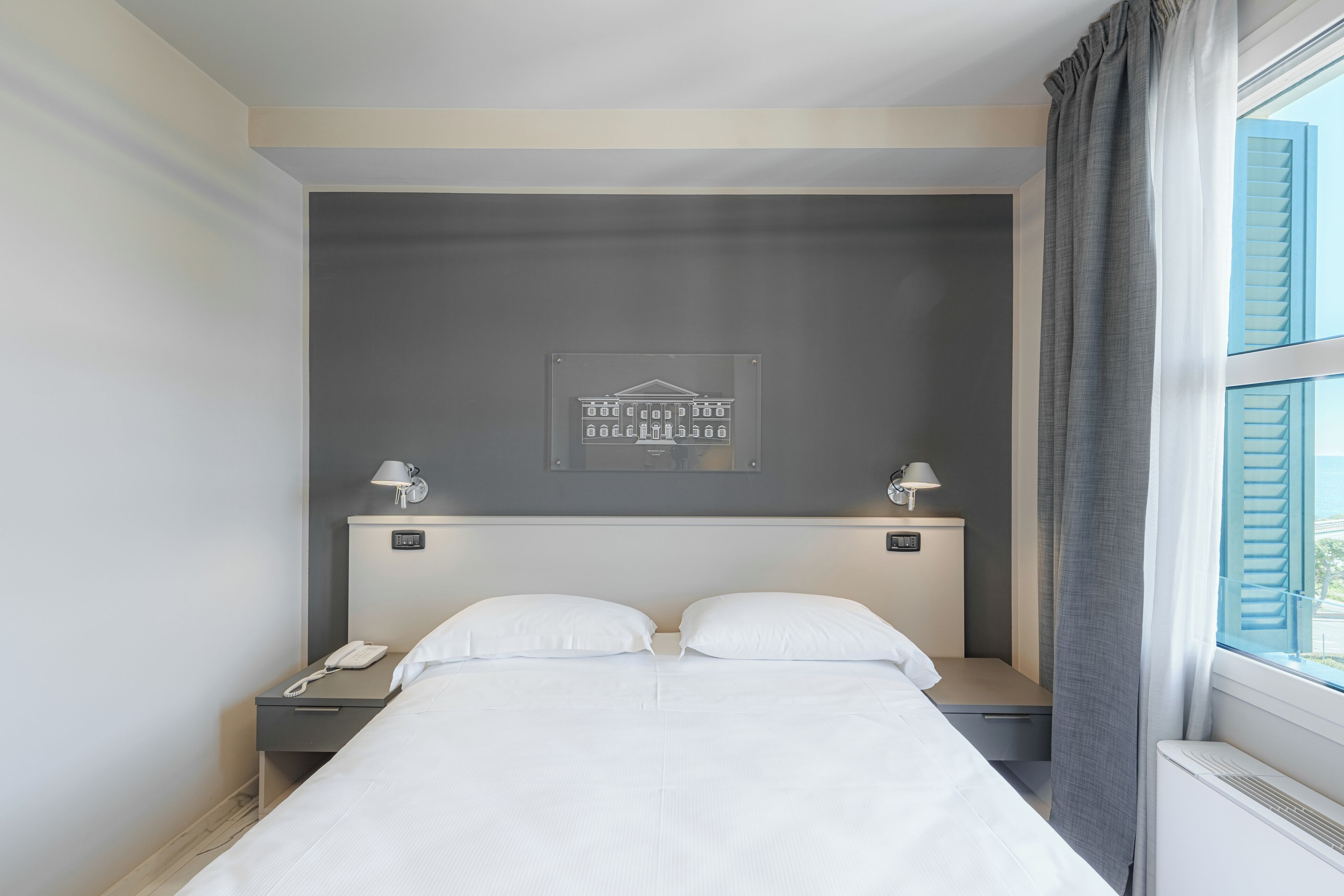 Una camera d'albergo con un letto, lampade da comodino, un comodino con un telefono, un quadro appeso al muro e una finestra con tende.