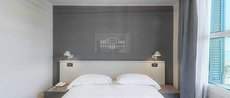 A hotel room with a bed, bedside lamps, a nightstand with a phone, a framed picture on the wall, and a window with curtains.
