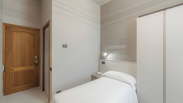 Habitación de hotel con puerta de madera, mesita de noche con lámpara, cama con ropa blanca de cama y armario.