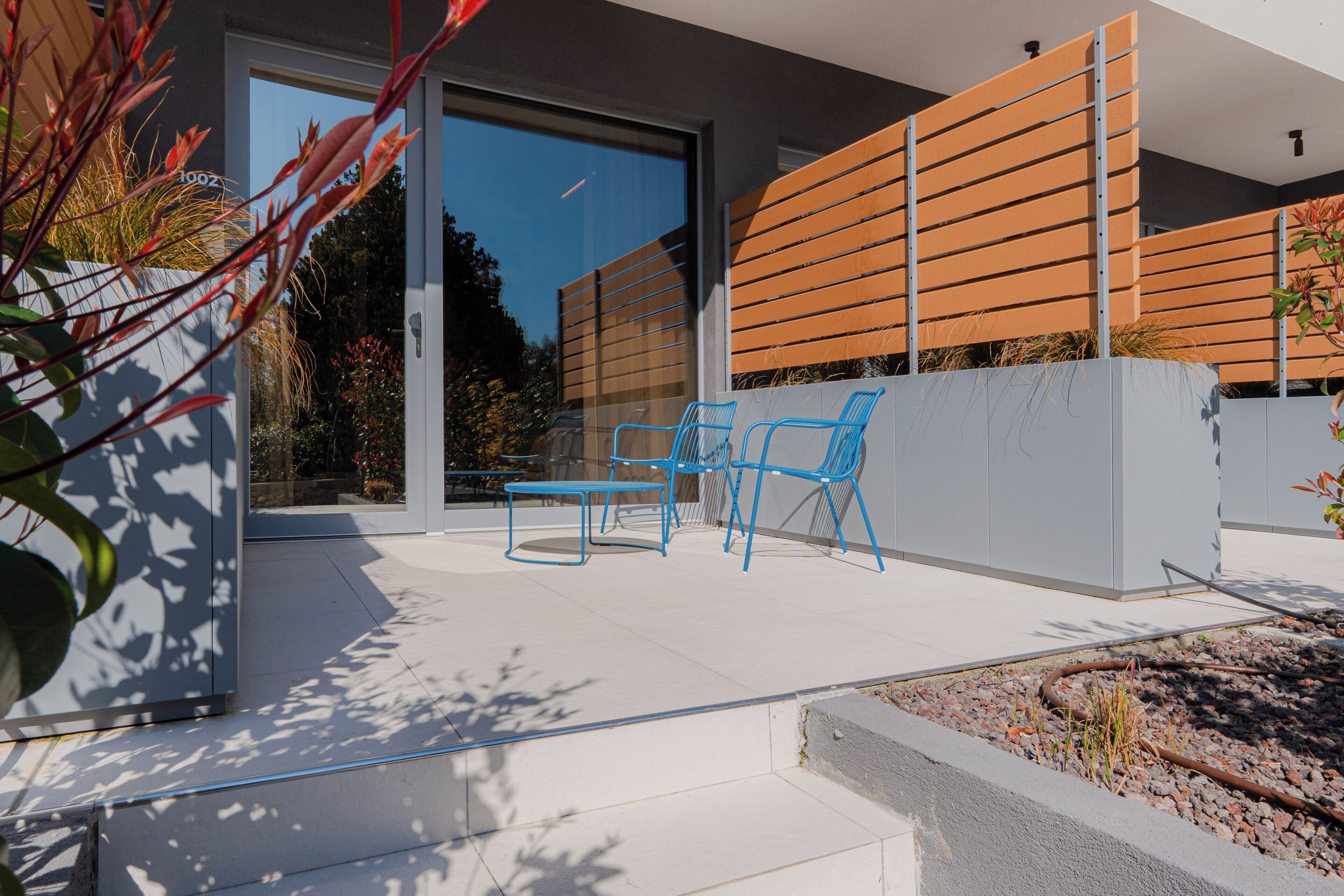 An outdoor patio with a modern design, featuring a seating area with chairs and a table, a glass door entrance, and a wooden slatted wall.