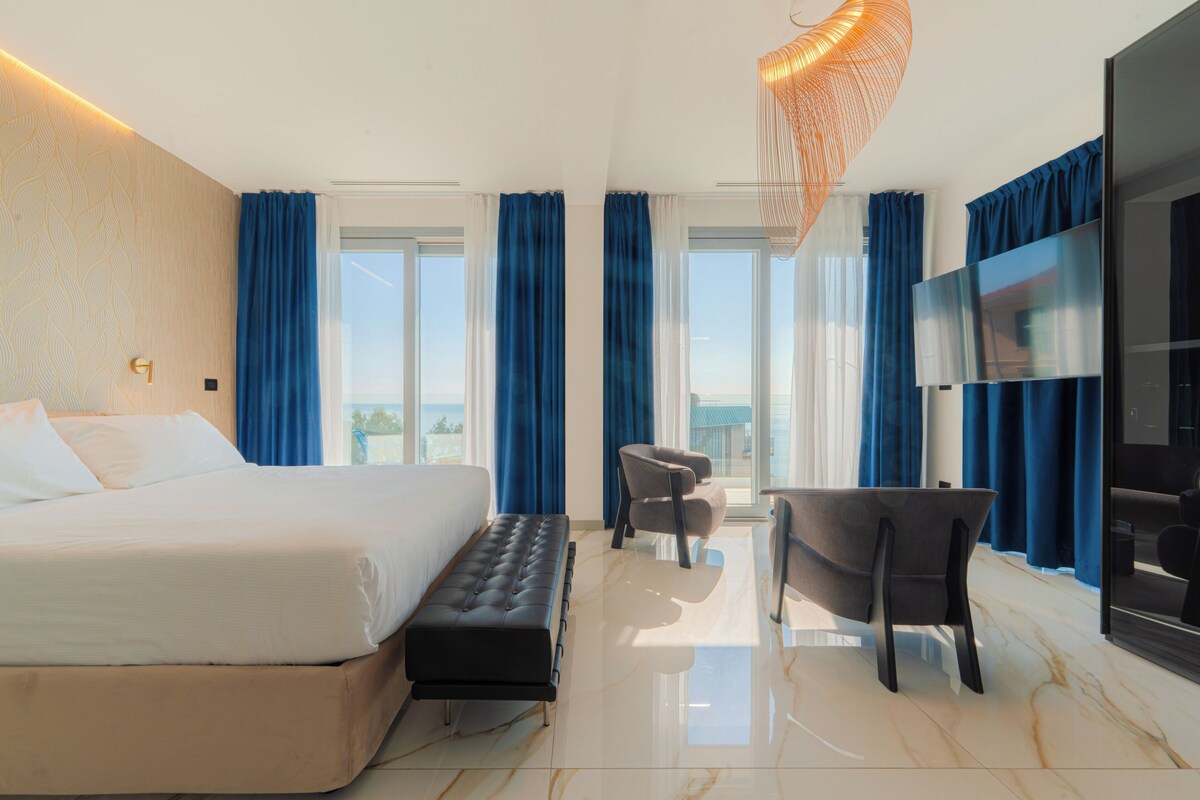 A modern hotel room with a large bed, a seating area, a flat-screen TV, and a view of the outdoors.