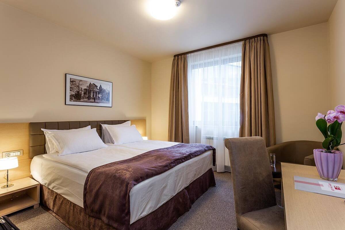 Una habitación de hotel con cama, escritorio, silla, lámpara, un florero con flores y un cuadro en la pared.