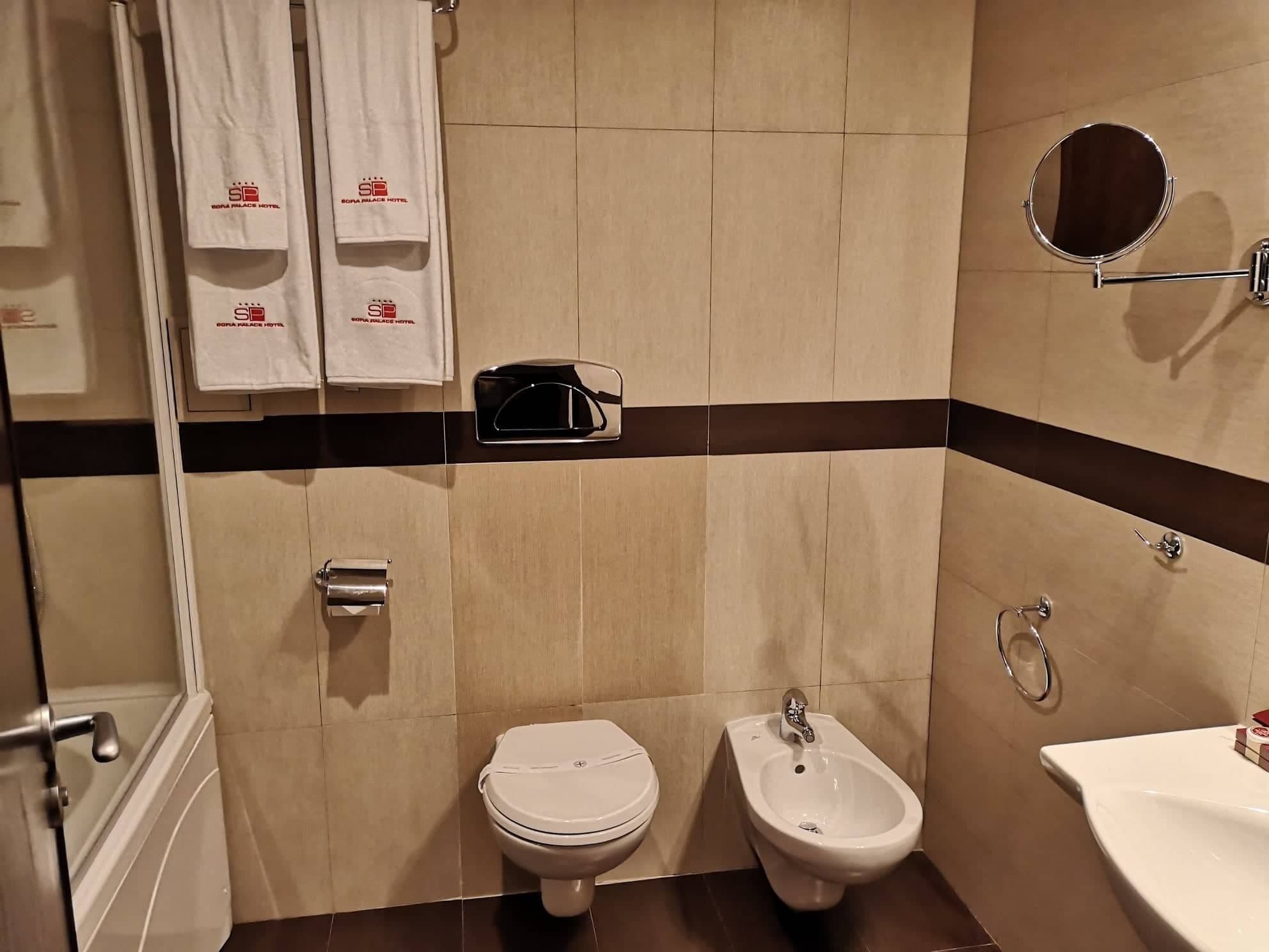 Casa de banho com sanita, bidé e lavatório, com parede em azulejo com faixa escura e toalheiro com toalhas da marca.
