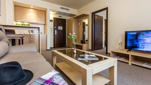 Quarto de hotel moderno com sofá, mesa de centro com livro e flores, televisão, área de cozinha e sinal de saída.