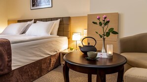 Quarto de hotel com uma cama, mesa de cabeceira com um conjunto de chá, um vaso com flores, uma vela acesa e um quadro na parede.