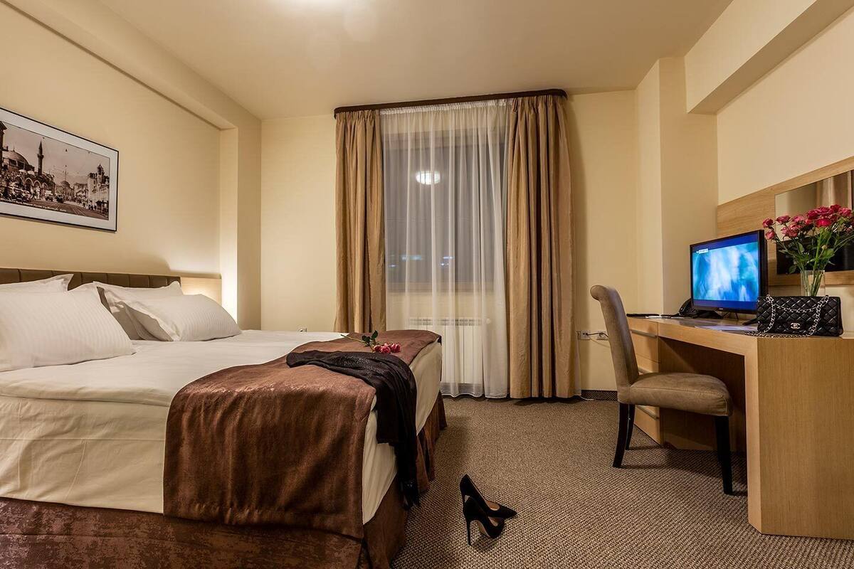 Habitación de hotel con una cama, un escritorio con computadora, una silla y un ventanal con cortinas.