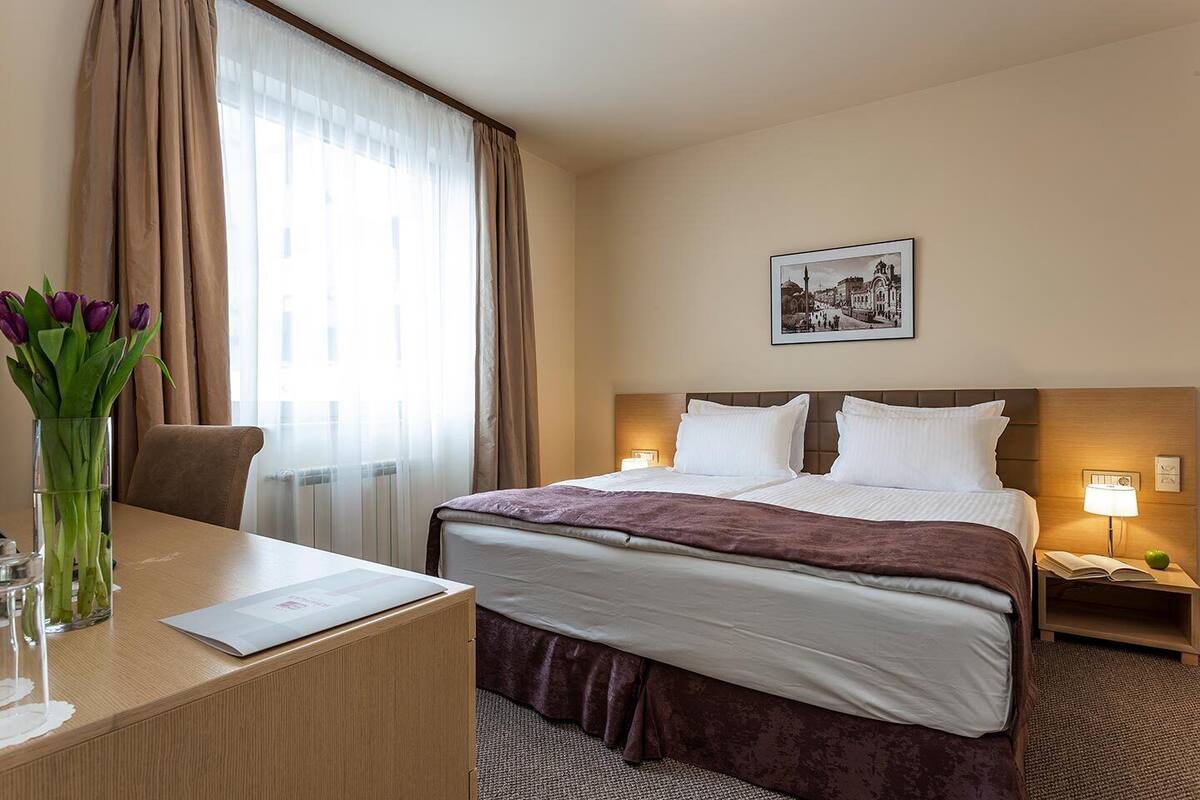 Habitación de hotel con cama, escritorio, silla, un jarrón con flores, un cuadro en la pared y una mesita de noche con lámpara.