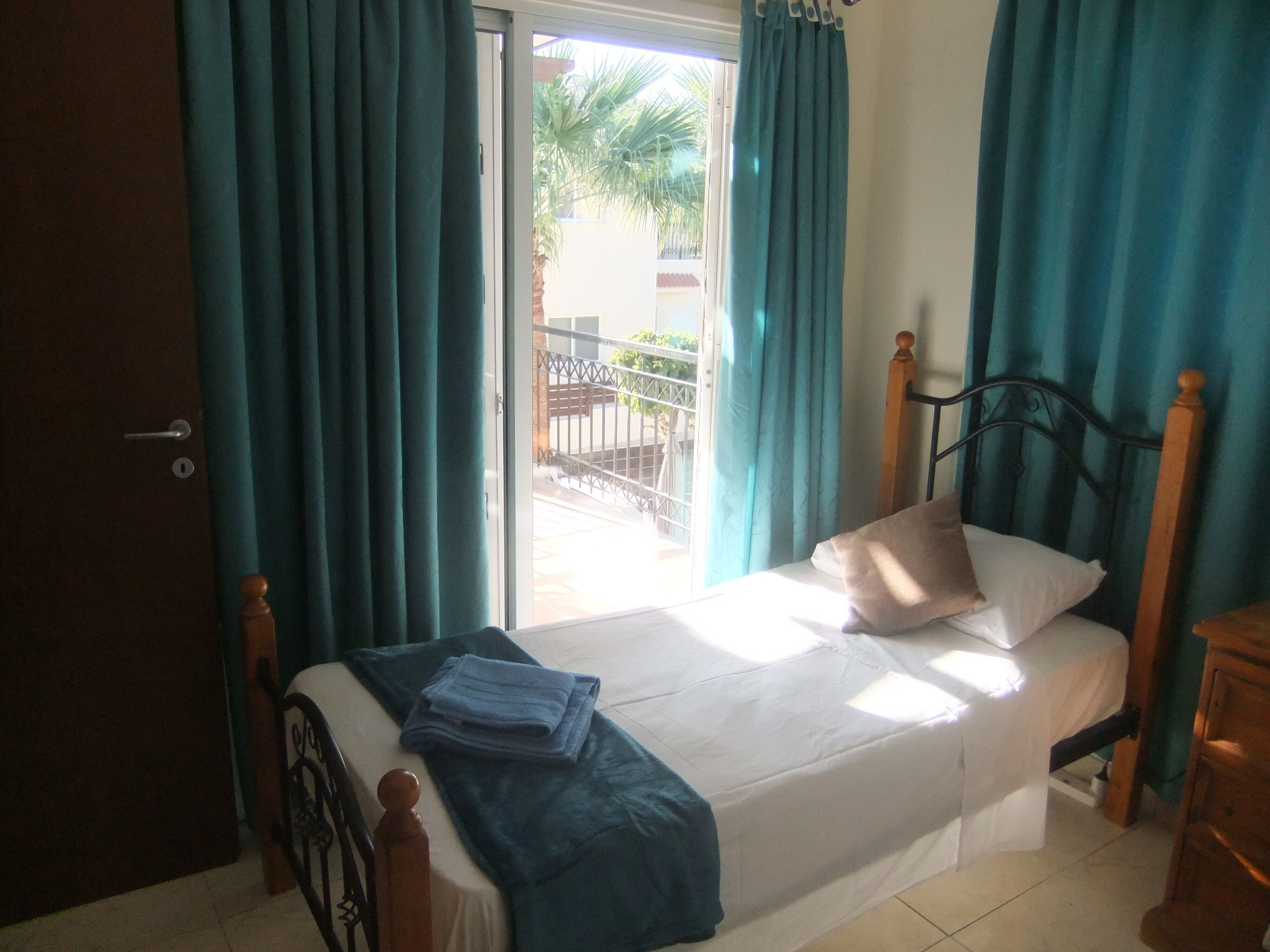 A room with a bed, a wooden headboard, a window with curtains, and a view of a balcony.