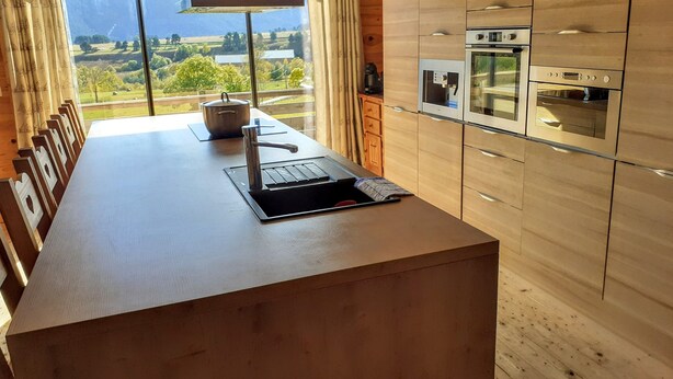 Une cuisine moderne avec des meubles en bois, un four encastré et une grande fenêtre offrant une vue panoramique sur les montagnes et la verdure.