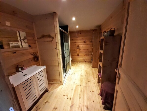 A wooden cabin interior with a wooden floor, a white sink, a glass shower door, and a wooden shelf with a basket.