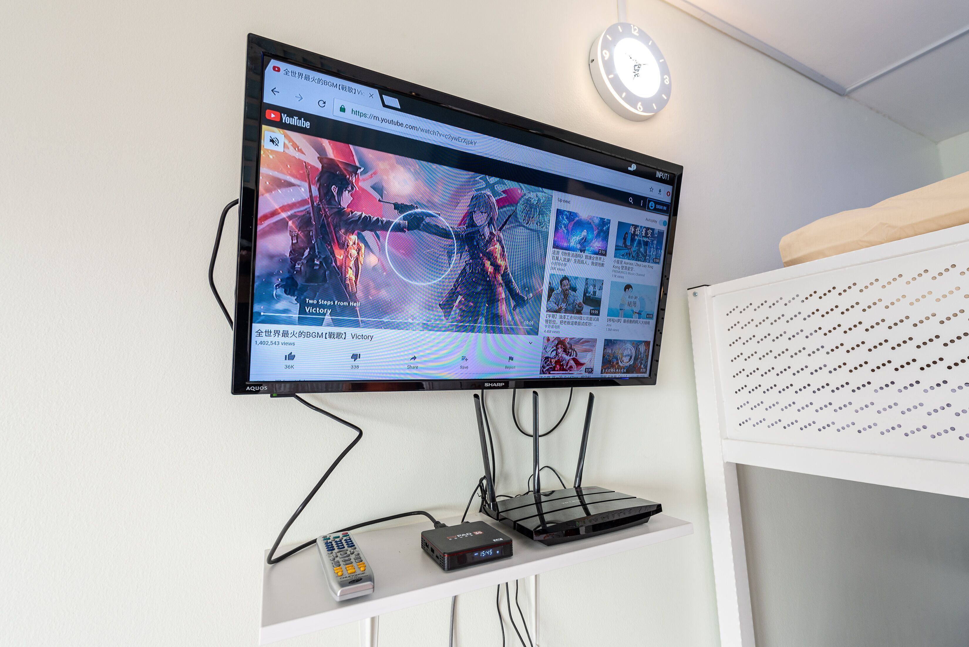 Un televisor de pantalla plana montado en la pared muestra un video de YouTube. Debajo del televisor, hay un estante blanco con un control remoto y un dispositivo electrónico negro.