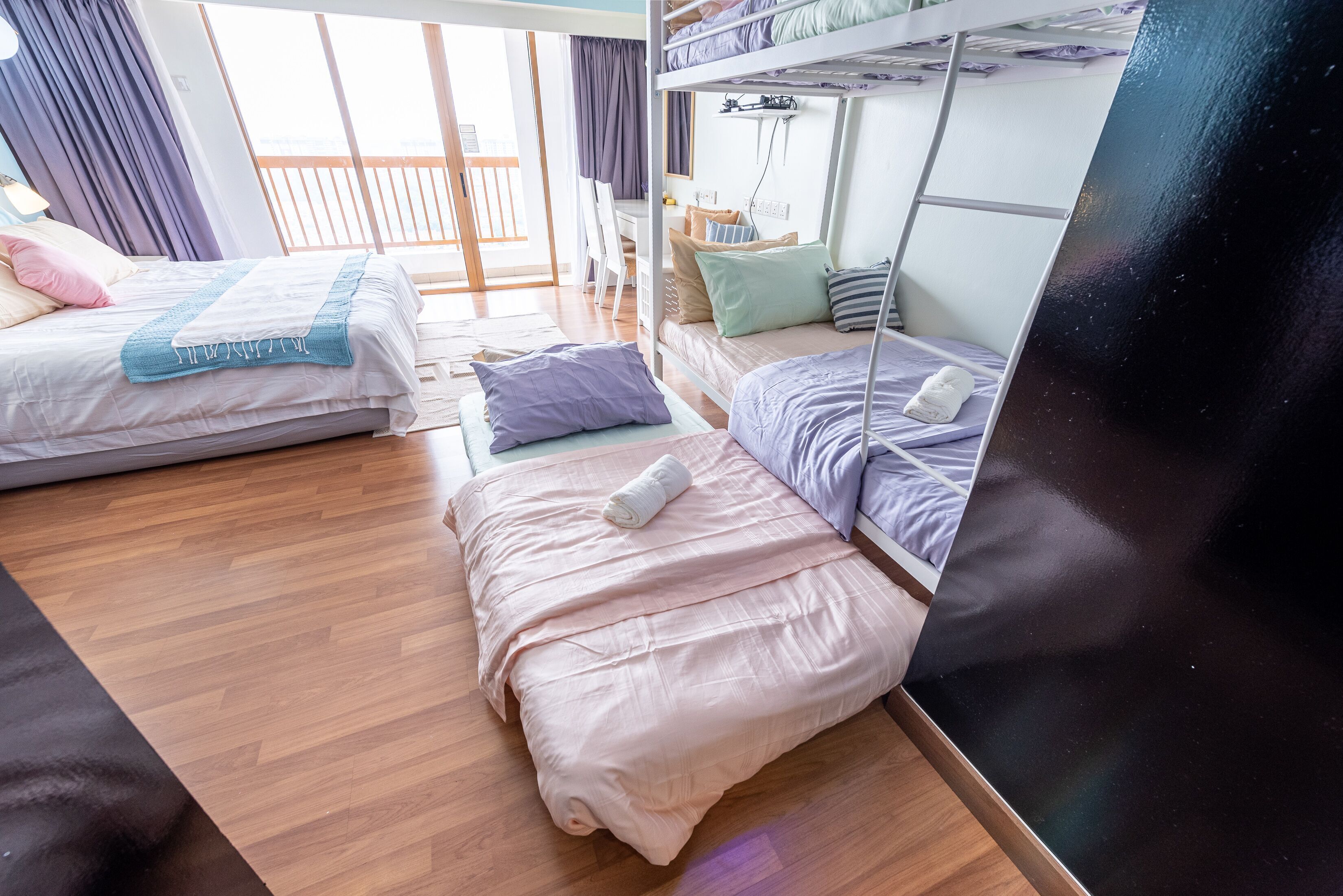 A room with two beds, wooden flooring, a desk, and a balcony with curtains.