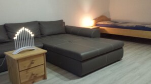 A modern bedroom with a grey sectional sofa, a wooden nightstand with drawers, a bedside table with a lamp, and a bed with a blue mattress. - Monteurzimmer & Ferienwohnung G. Klement (Herrieden)