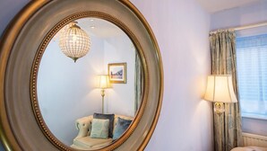 A room with a large oval mirror, a sofa with cushions, a lamp, and a window with curtains. - Henley Hotel (Henley-in-Arden)