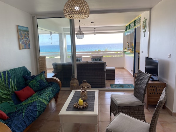 A living room with a sofa, a coffee table, and a TV. There is a balcony with a view of the ocean.