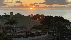A coastal sunset view with a house featuring a green roof and a white picket fence. - Magnifique appartement vue mer 180° - Hauteur anse Des Rochers - Avec Cuve Eau (Saint-François)