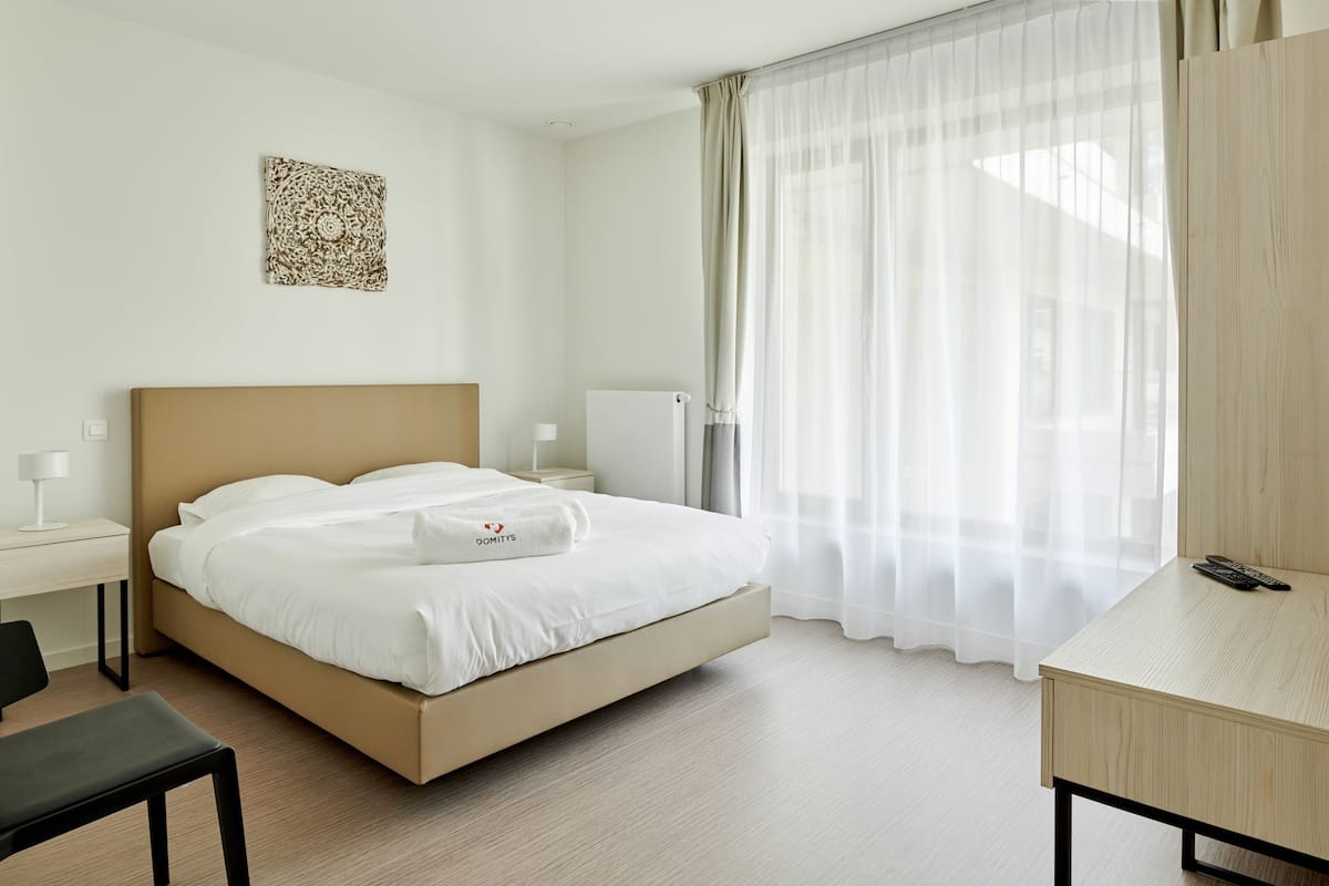 A hotel room with a bed, a desk, a chair, a bedside table, and a window with sheer curtains.