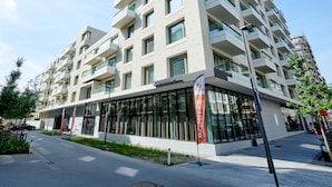 A modern multi-story building with a glass facade, a visible entrance, and a flag indicating a service area. - Domitys Emerald Haven (Antwerp)