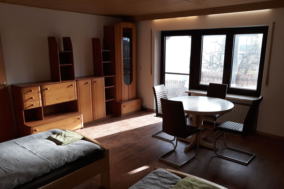 Quarto com cama, escrivaninha, cadeira, mesa e janela com vista para edifícios.
