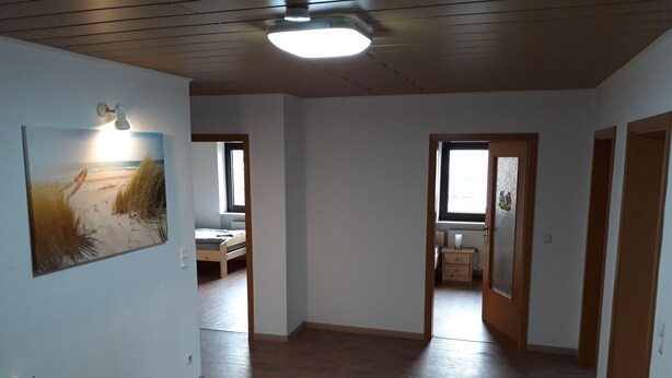 A spacious hallway with wooden floors and white walls, leading to a bedroom with a bed and a window.