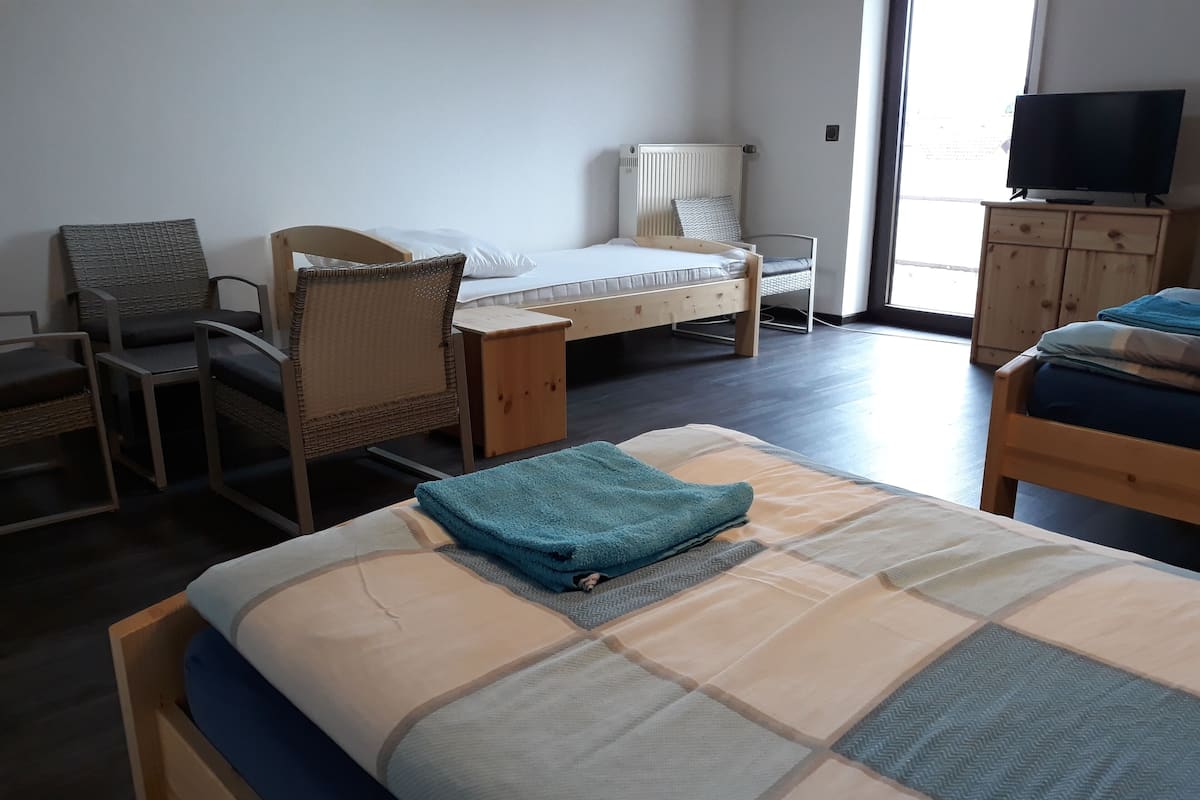 Quarto com cama, duas cadeiras, escrivaninha, televisão e janela.
